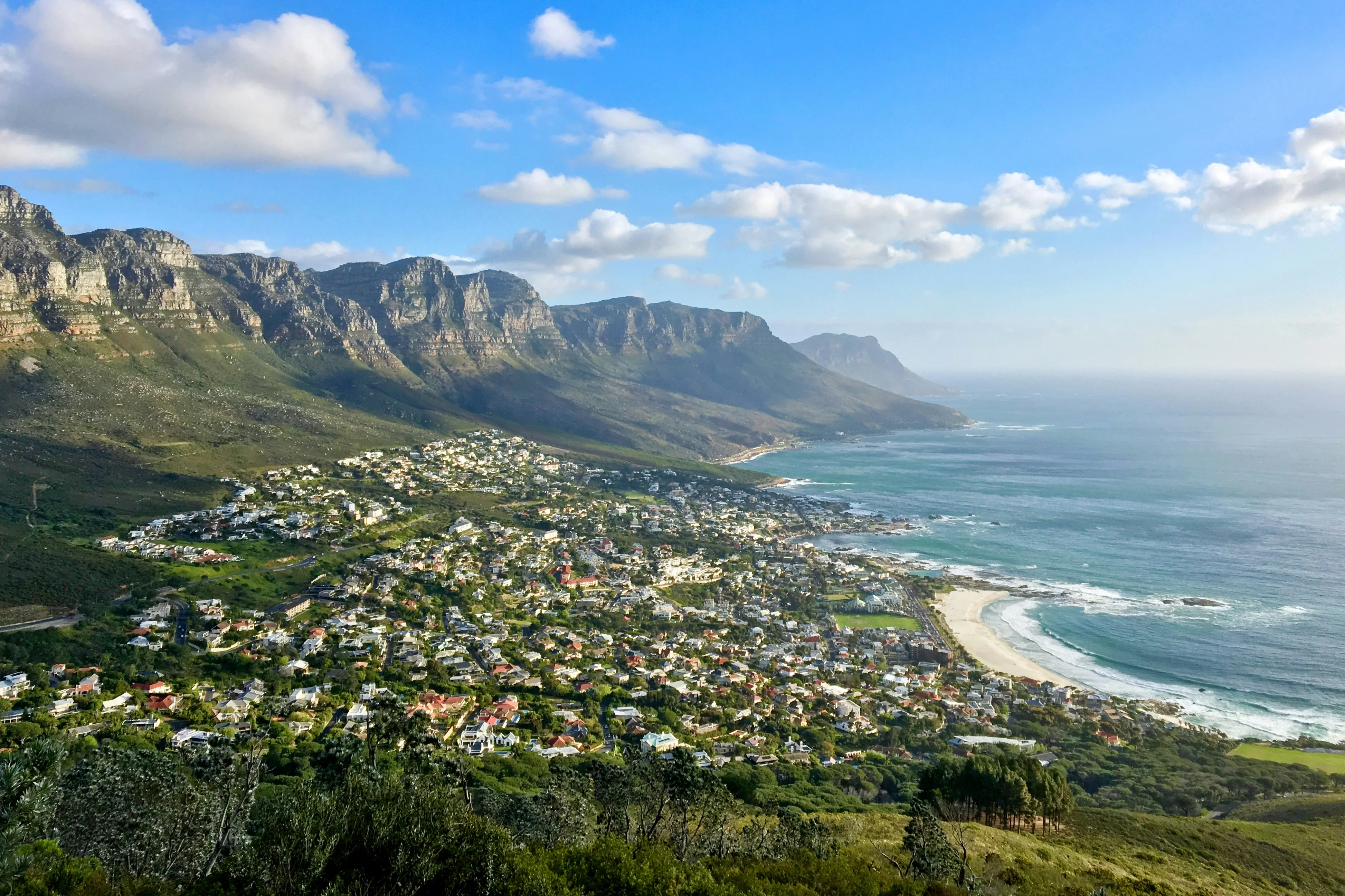 Aerial view of Camps Bay, Cape Town, showing luxury homes and the Atlantic Ocean against the Twelve Apostles mountain range.
